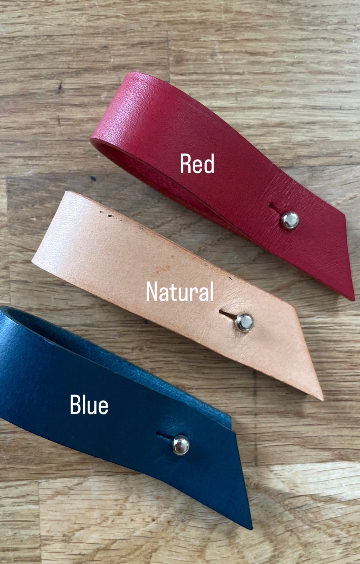 Three leather strap pieces in red, natural tan, and blue, each with a silver metal rivet, laid out on a wooden surface with text labels 'Red', 'Natural', and 'Blue'.