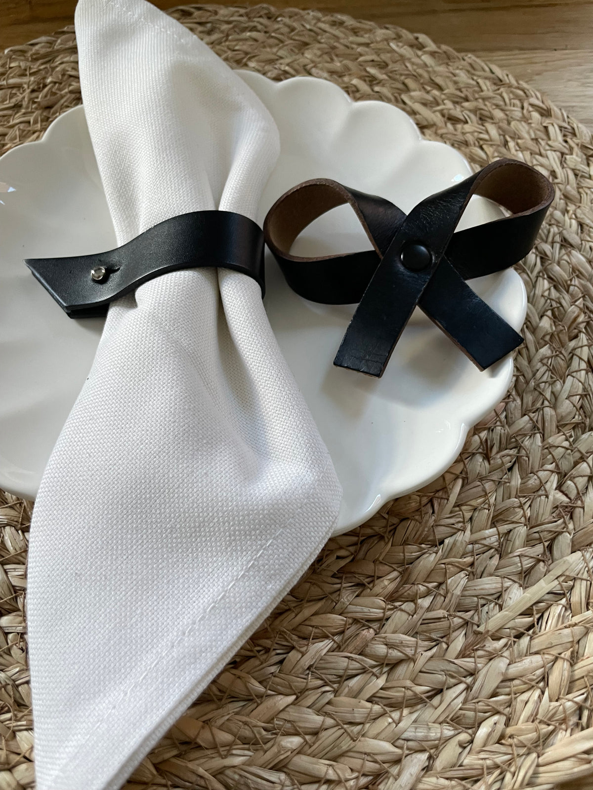 Two black leather napkin rings, one holding a white napkin on a white scalloped plate and the other resting beside it, set on a woven placemat.